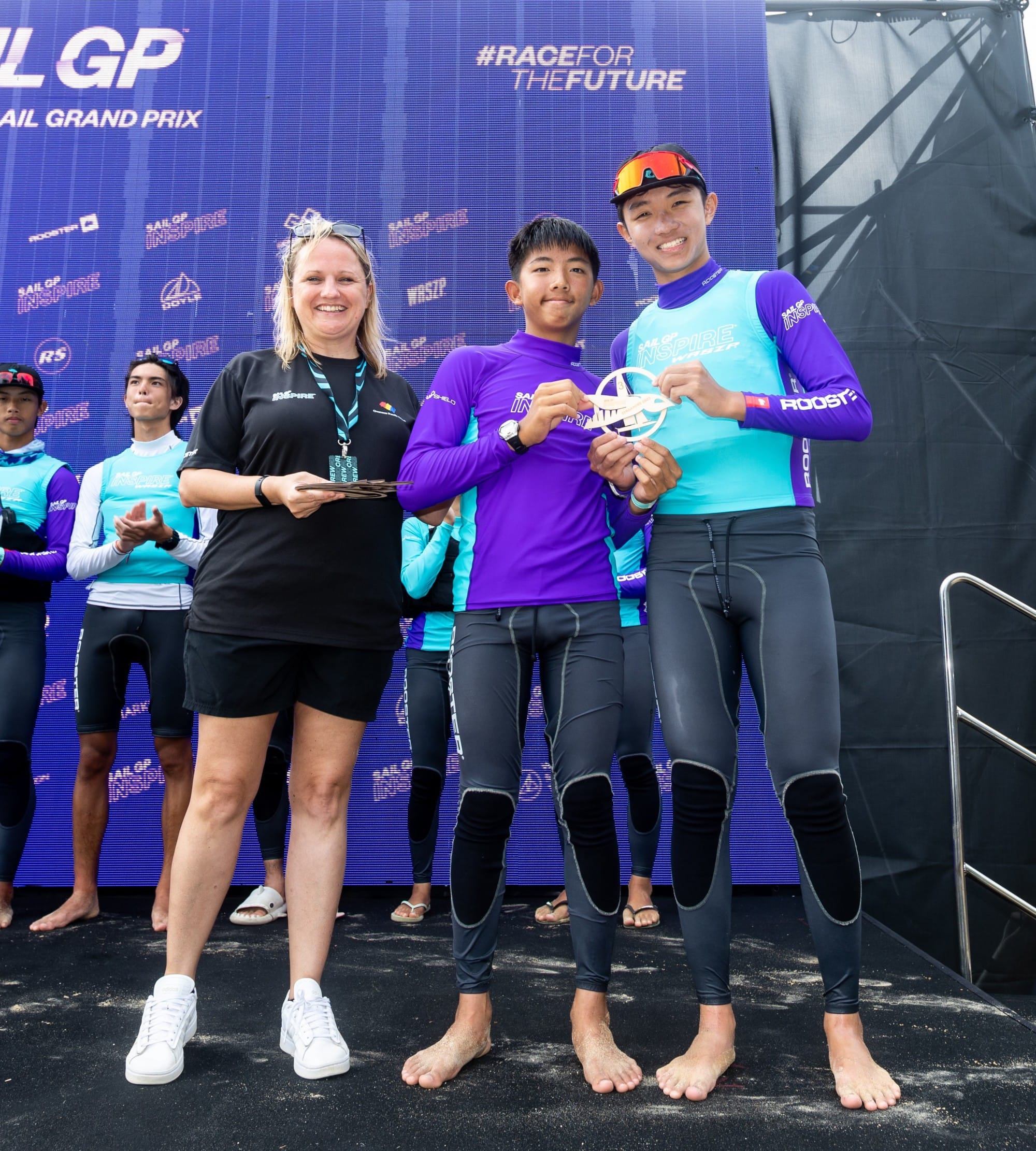 Three people smile at a Sail Grand Prix event, two young sailors in purple and teal holding a trophy-style award.